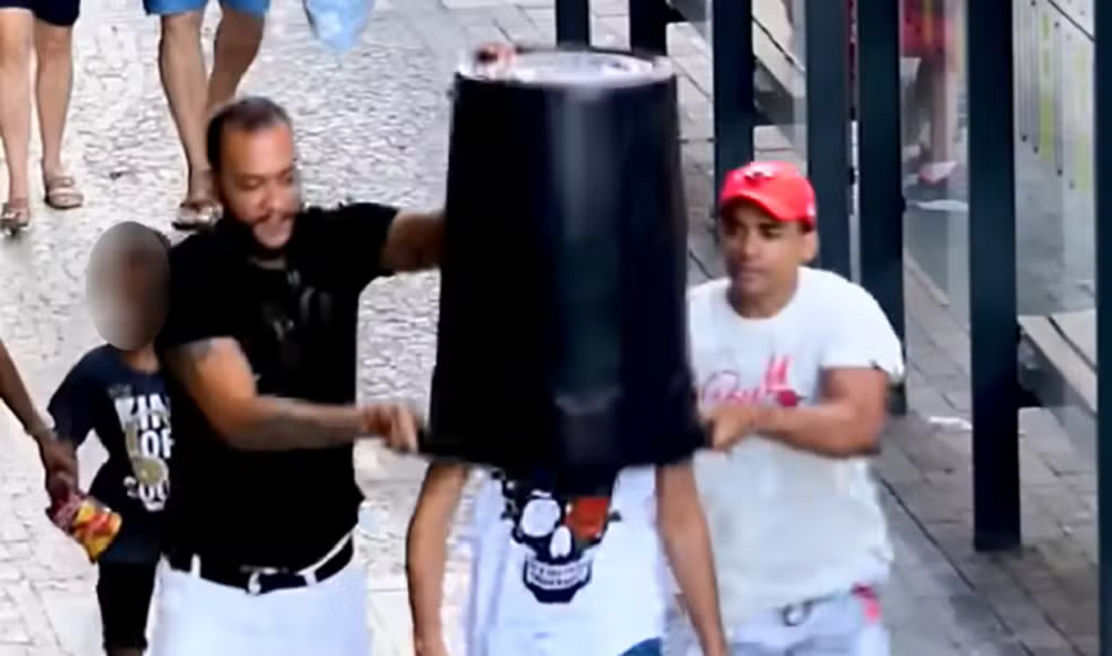 Dois homens colocando um balde na cabeça de outro homem durante pegadinha