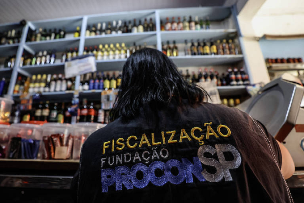 Agente fiscaliza bebidas em estabelecimento em São Paulo, de costas, com garrafas no fundo da foto