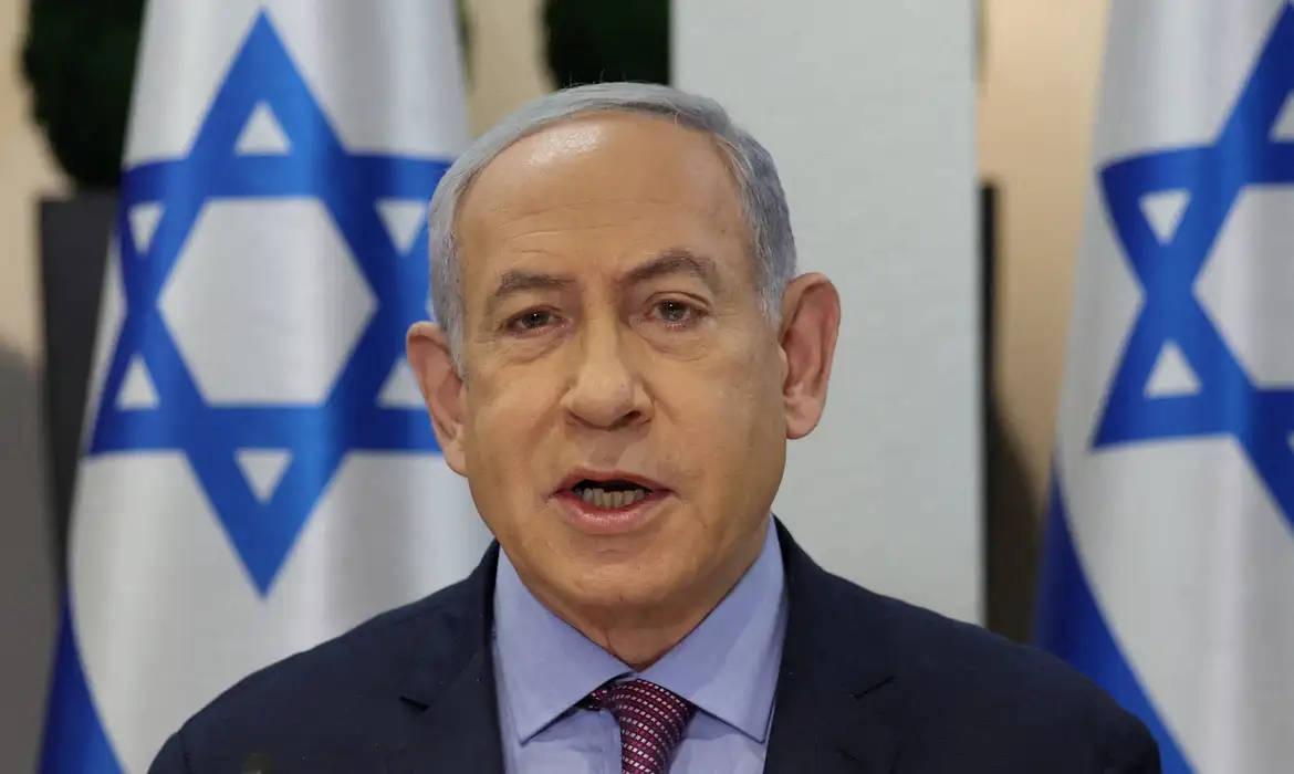 Benjamin Netanyahu falando, sério, com bandeiras de Israel ao fundo