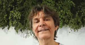 cantor e compositor Lô Borges sorrindo, de camiseta cinza, olhando para a câmera, com plantas ao fundo