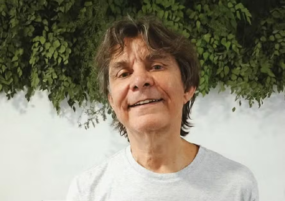 cantor e compositor Lô Borges sorrindo, de camiseta cinza, olhando para a câmera, com plantas ao fundo