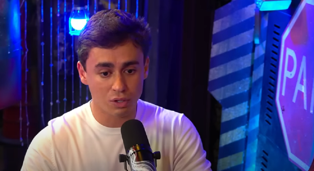 O deputado federal Nikolas Ferreira (PL) falando em microfone, sério, de camiseta branca, sem olhar para a câmera