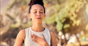 Mulher respirando de cabelo preso, camiseta branca e olhos fechados