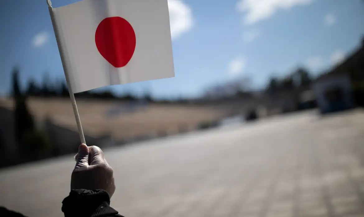 Mão com luva segurando pequena bandeira do Japão ao ar livre em dia ensolarado