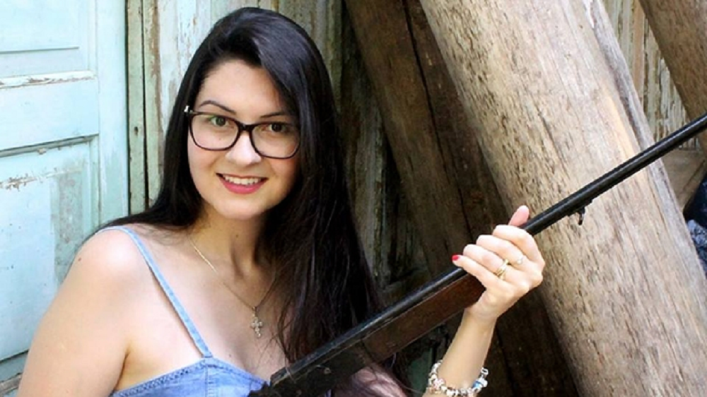 Ana Campagnolo de óculos, sorrindo e segurando arma