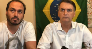 O vereador Carlos Bolsonaro (PL-RJ) e o ex-presidente Jair Bolsonaro (PL) sentados lado a lado, sérios, de roupas brancas, sem olhar para a câmera