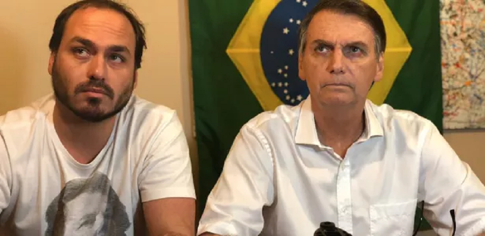 O vereador Carlos Bolsonaro (PL-RJ) e o ex-presidente Jair Bolsonaro (PL) sentados lado a lado, sérios, de roupas brancas, sem olhar para a câmera