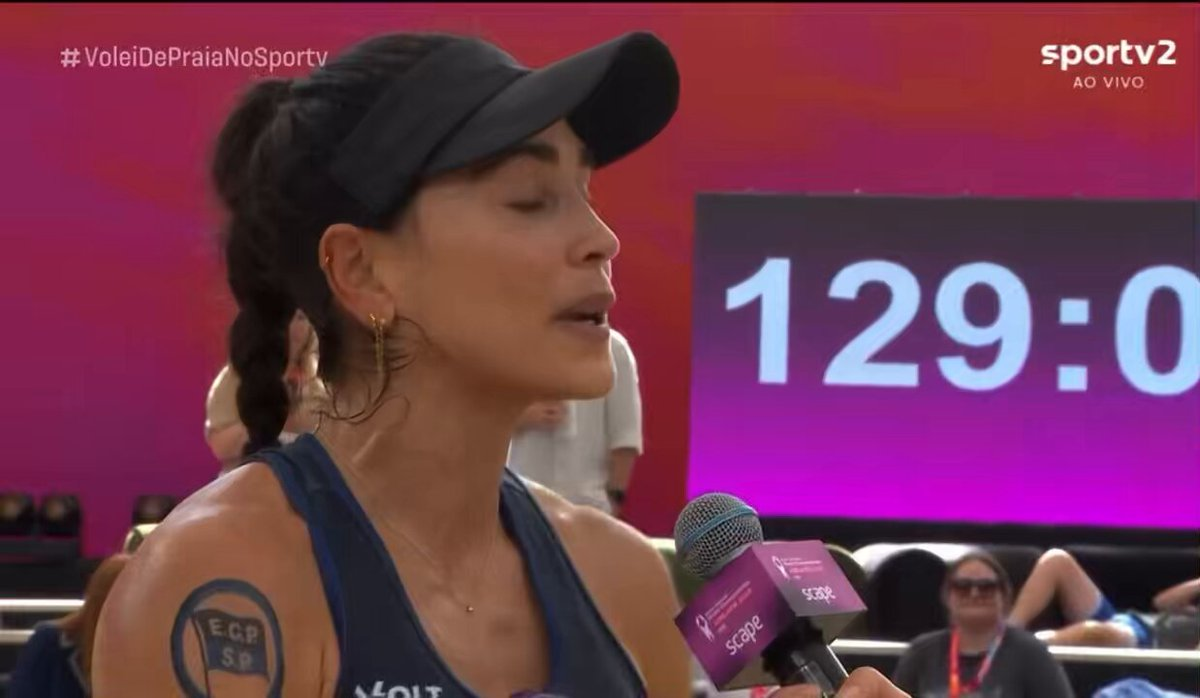 Carol Solberg falando em microfone, de viseira, de olhos fechados, de perfil