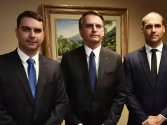 Flávio, Jair e Eduardo Bolsonaro em pé, sérios, lado a lado, olhando para a câmera, com quadro na parede ao fundo