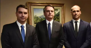 Flávio, Jair e Eduardo Bolsonaro em pé, sérios, lado a lado, olhando para a câmera, com quadro na parede ao fundo