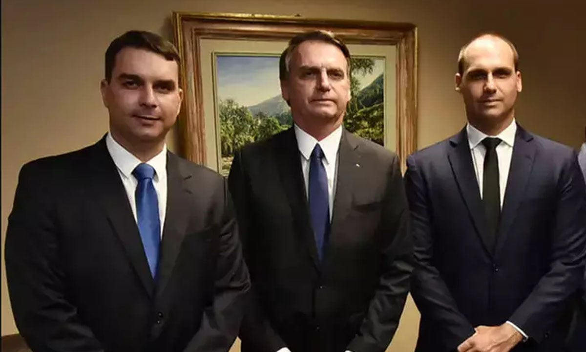 Flávio, Jair e Eduardo Bolsonaro em pé, sérios, lado a lado, olhando para a câmera, com quadro na parede ao fundo