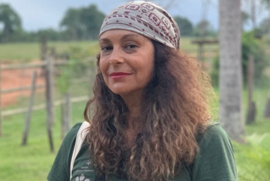 A atriz Giovanna Goldfarb de bandana e cabelo solto, maquiada, olhando séria para a câmera, ao ar livre