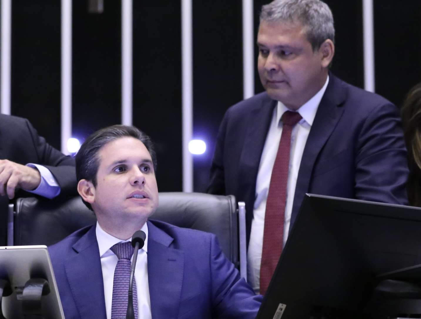Os deputados Hugo Motta e Lindbergh Farias, sérios, o primeiro sentado e o segundo ao lado, em pé