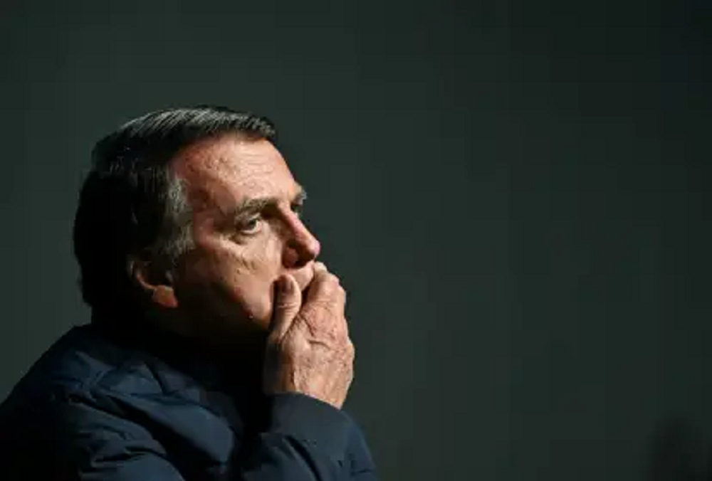 Jair Bolsonaro no canto esquerdo de foto, de perfil, sério, com mão na boca