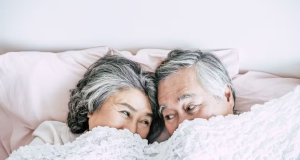 Casal de idosos deitado embaixo de cobertor e se olhando, apenas com uma parte do rosto descoberta