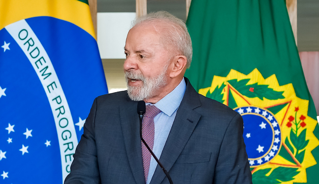 Lula olhando para o lado e falando, com bandeiras do Brasil ao fundo