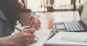 Pessoa escrevendo perto de notebook, com copo de café