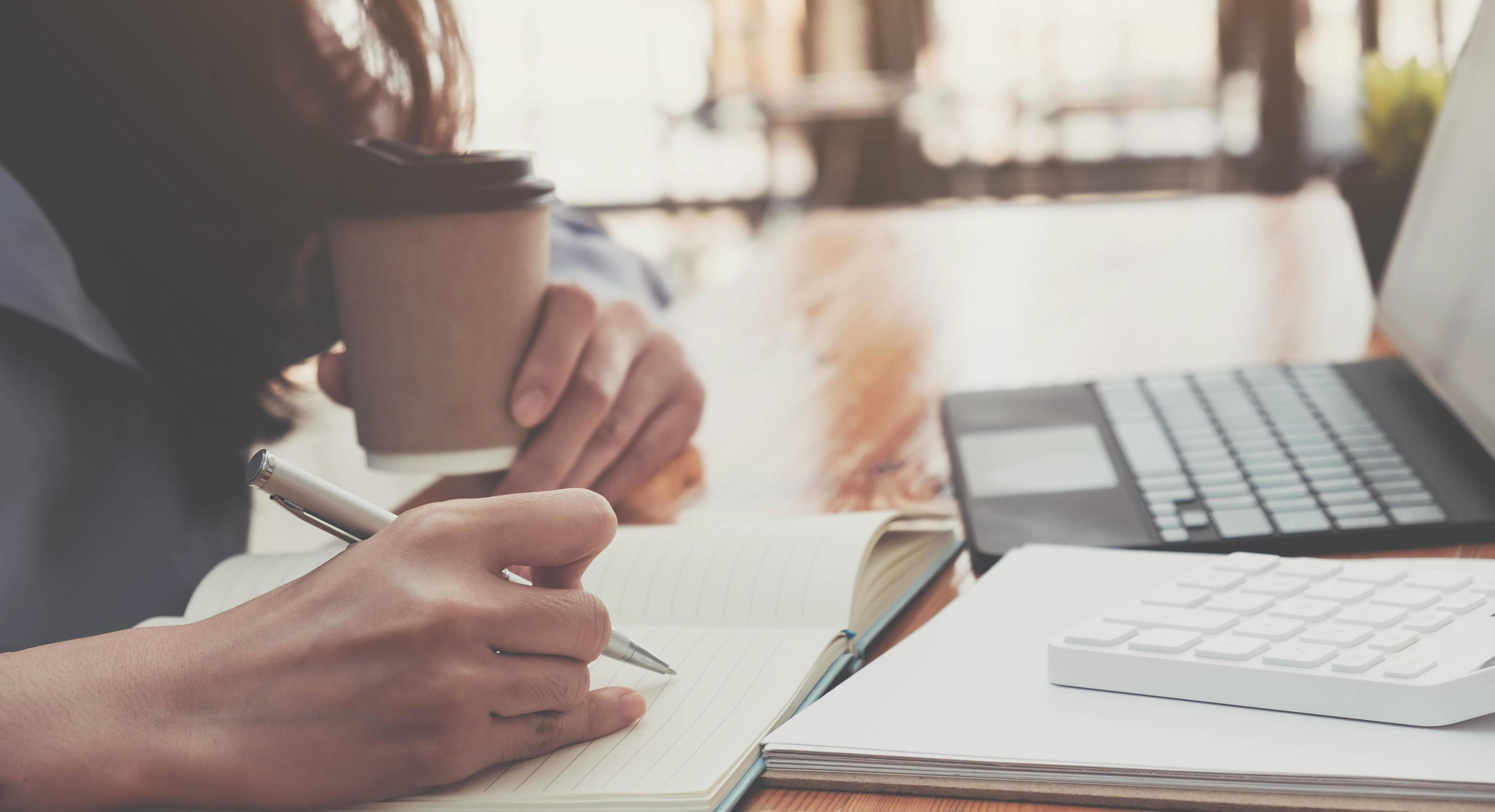 Pessoa escrevendo perto de notebook, com copo de café