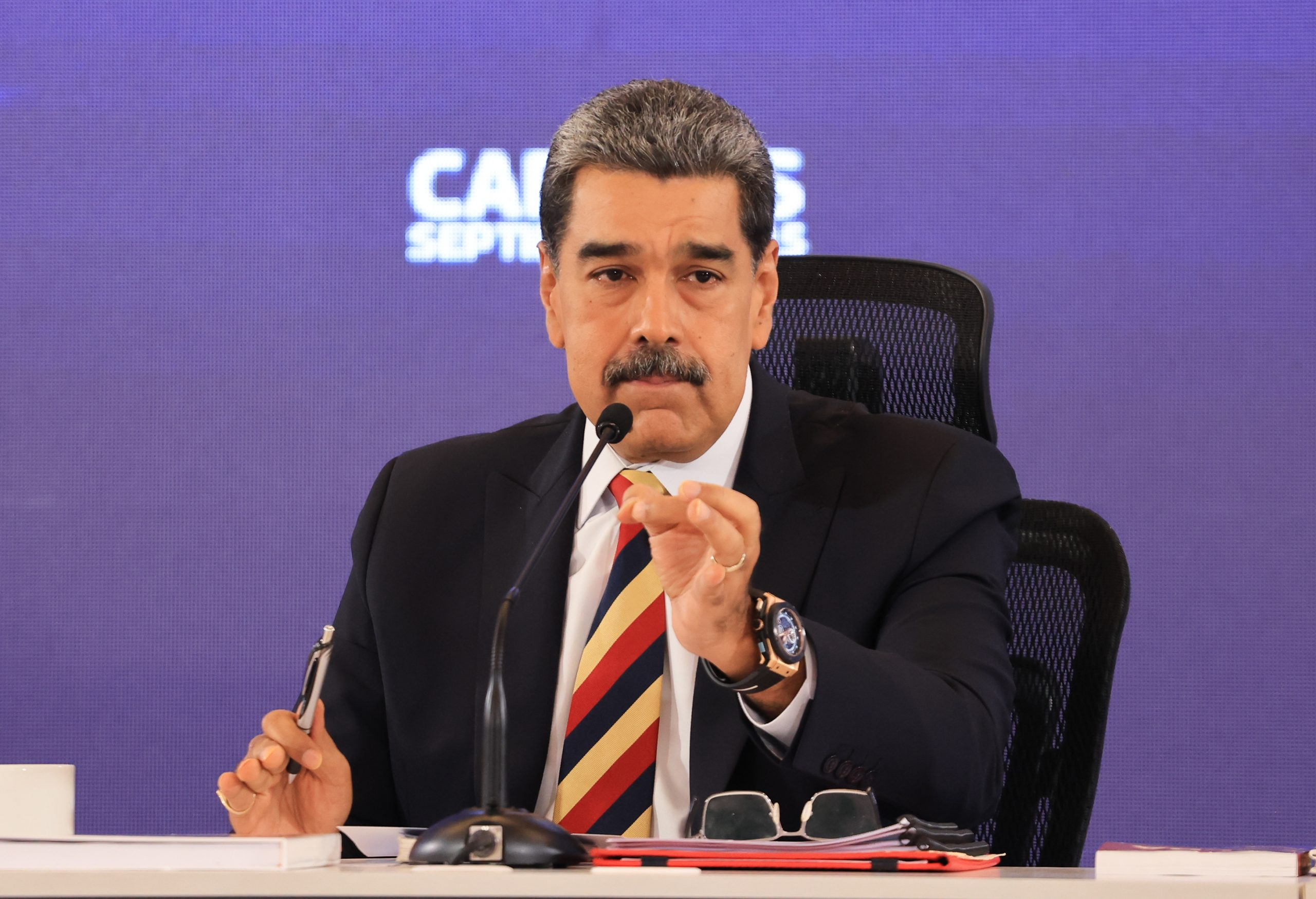 Nicolás Maduro falando em microfone e gesticulando, sério, sem olhar para a câmera, sentado
