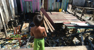 Menino sem camisa, de bermuda verde, de costas, em pé, em comunidade