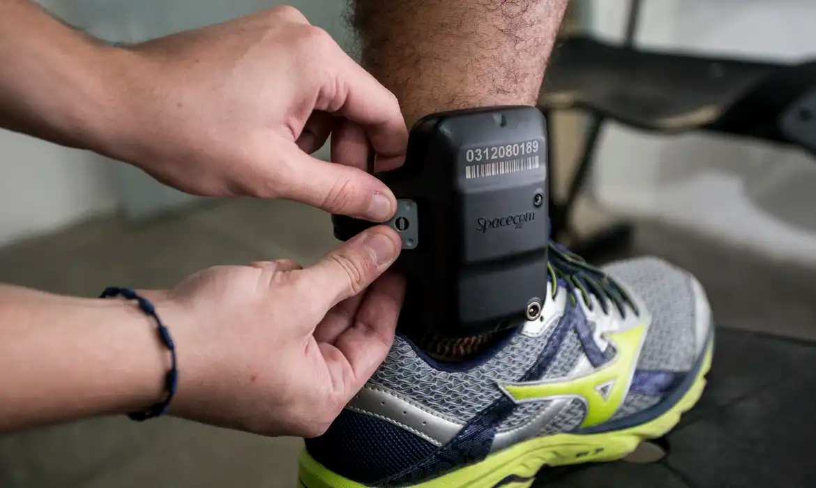 Perna com tornozeleira eletrônica sendo ajustada por duas mãos, pessoa calçando tênis
