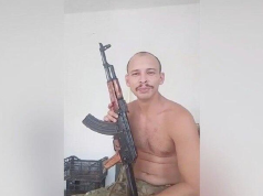 Philippe Marques Pinto no meio de montagem, sem camisa, segurando fuzil