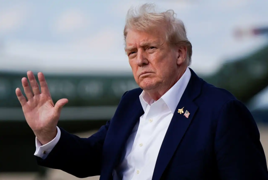 O presidente americano Donald Trump sério, sem olhar para a câmera, acenando ao ar livre