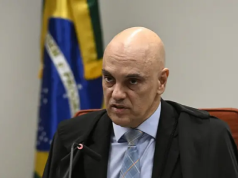 Alexandre de Moraes falando em microfone, com bandeira do Brasil ao fundo, sério, sem olhar para a câmera