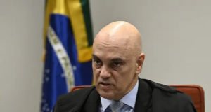 Alexandre de Moraes falando em microfone, com bandeira do Brasil ao fundo, sério, sem olhar para a câmera