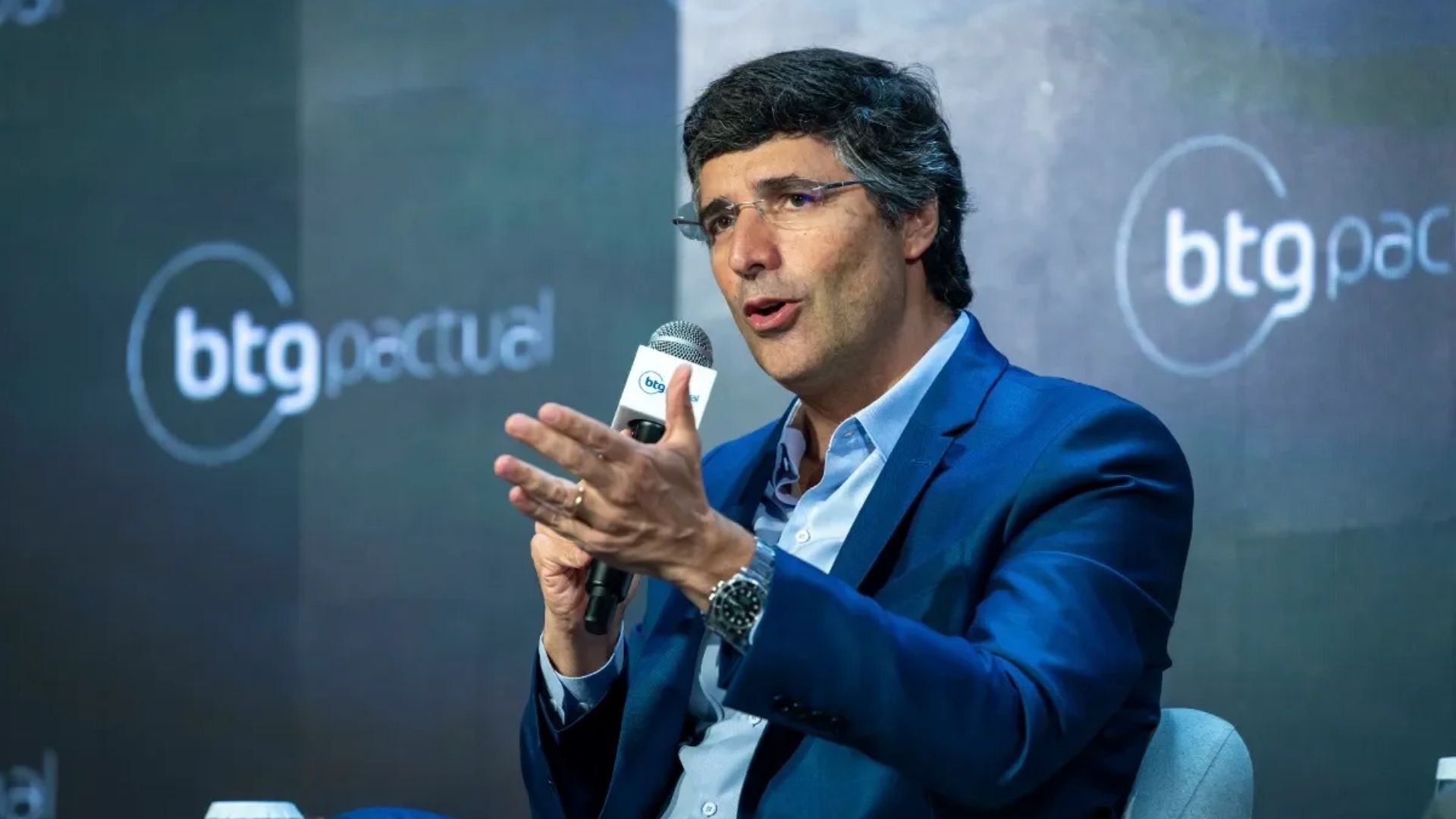 O empresário André Esteves, do BTG Pactual, sentado, falando em microfone e gesticulando