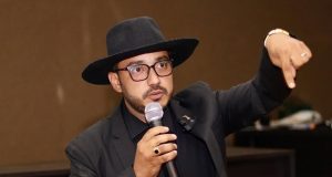 O poeta, advogado e influenciador Dudu Morais, de roupa e chapéu pretos, falando em microfone e apontando pra baixo, sério, de óculos de grau, sem olhar para a câmera