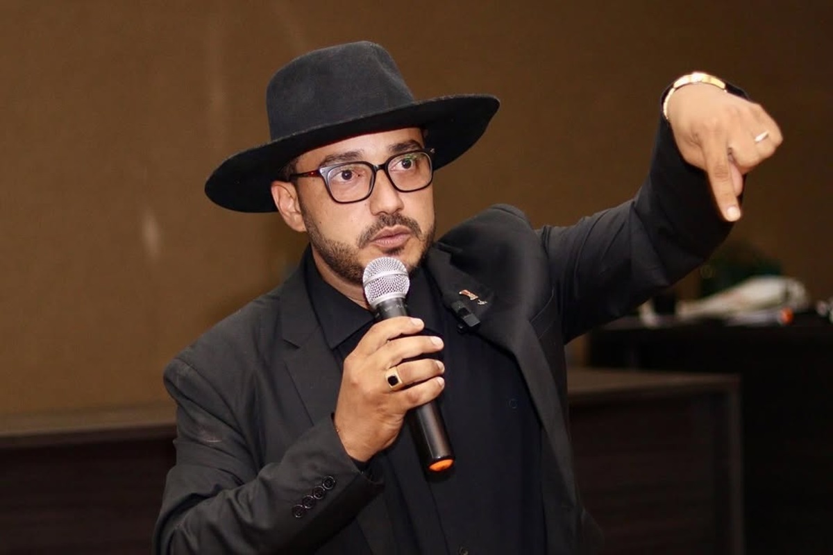 O poeta, advogado e influenciador Dudu Morais, de roupa e chapéu pretos, falando em microfone e apontando pra baixo, sério, de óculos de grau, sem olhar para a câmera