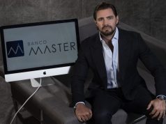 Daniel Vorcaro, dono do Banco Master, sentado ao lado de computador com a logo do banco, olhando para a câmera, sério