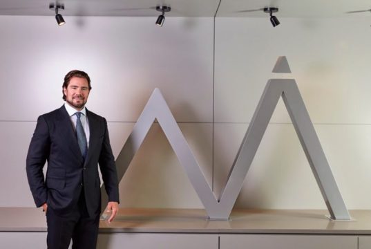 O dono do Banco Master, Daniel Vorcaro, em pé, sorrindo, perto de símbolo do banco