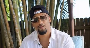 O influenciador bolsonarista Alex Oliveira de boné para trás, óculos escuros e camisa social branca, sério, falando para a câmera