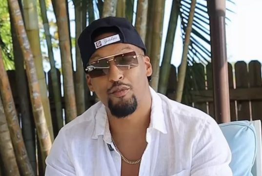 O influenciador bolsonarista Alex Oliveira de boné para trás, óculos escuros e camisa social branca, sério, falando para a câmera