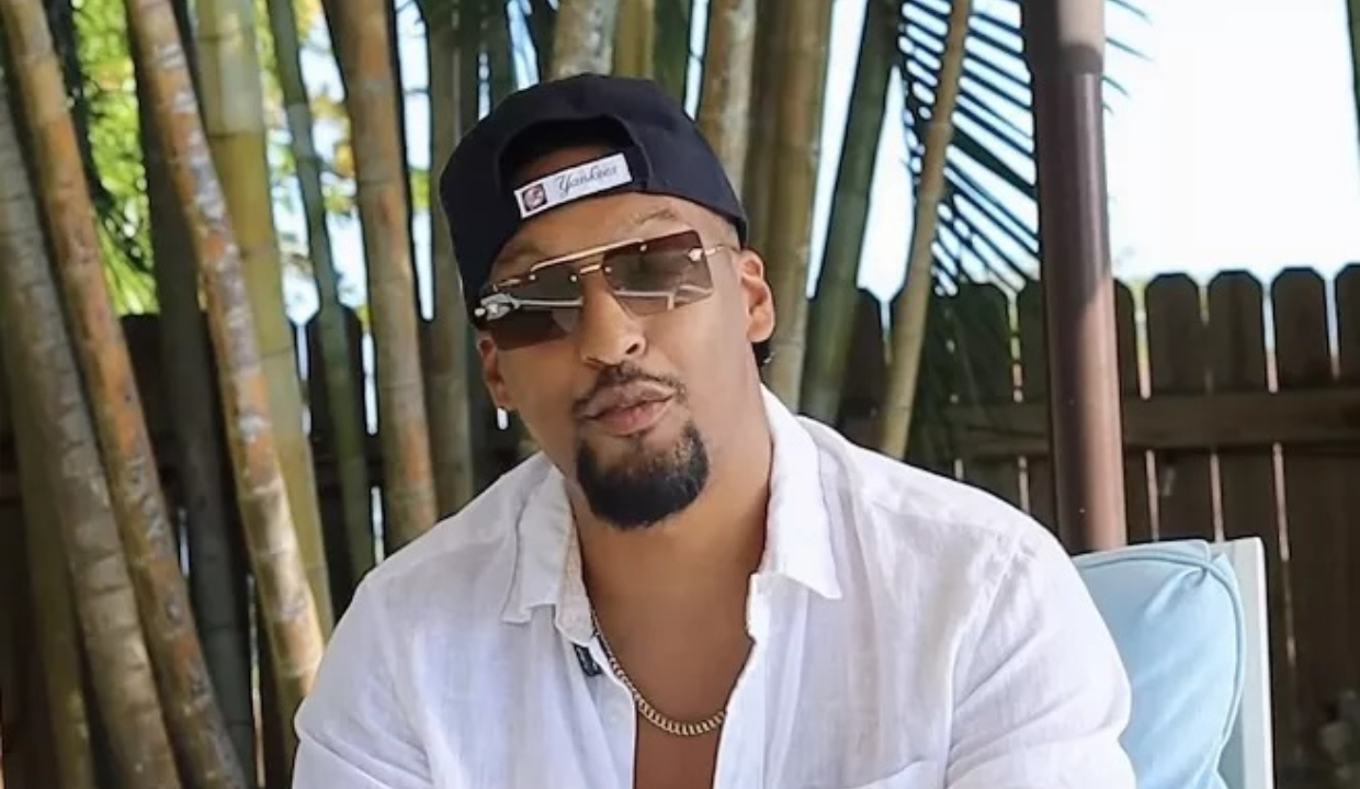 O influenciador bolsonarista Alex Oliveira de boné para trás, óculos escuros e camisa social branca, sério, falando para a câmera