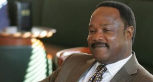 Isiah Whitlock Jr. com sorriso tímido em The Wire, sem olhar para a câmera, de roupa social marrom