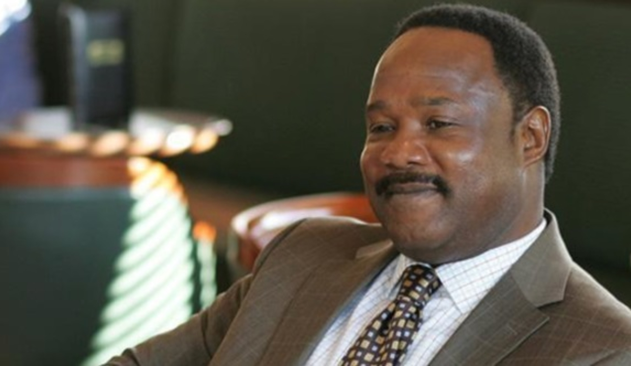 Isiah Whitlock Jr. com sorriso tímido em The Wire, sem olhar para a câmera, de roupa social marrom