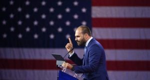 ex-deputado Eduardo Bolsonaro (PL-SP) em pé, falando e apontando, com bandeira dos EUA ao fundo