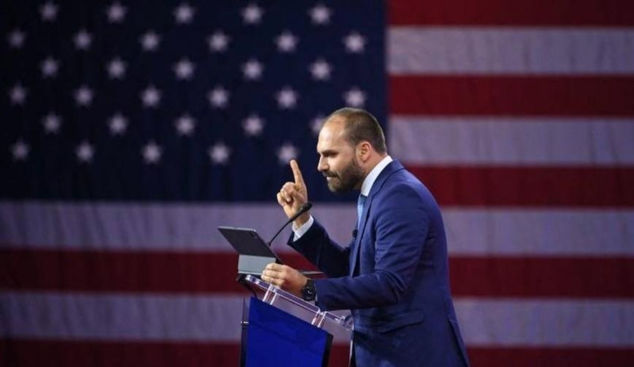 ex-deputado Eduardo Bolsonaro (PL-SP) em pé, falando e apontando, com bandeira dos EUA ao fundo
