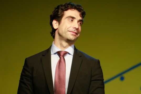 Gabriel Galípolo de terno e gravata, olhando para o lado com sorriso tímido