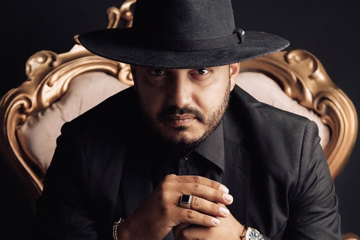 Dudu Morais de roupa e chapéu pretos, sério, sentado e olhando para a câmera