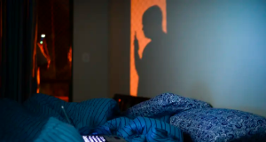 Pessoa em pé, mexendo no celular, no escuro, com cama bagunçada e notebook aberto em cima da cama