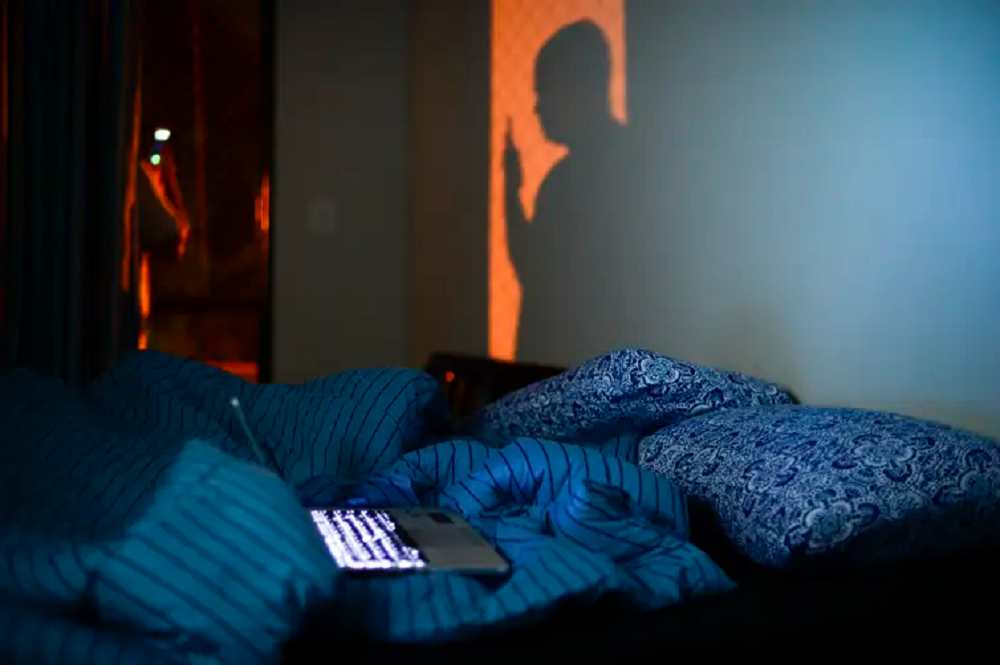 Pessoa em pé, mexendo no celular, no escuro, com cama bagunçada e notebook aberto em cima da cama