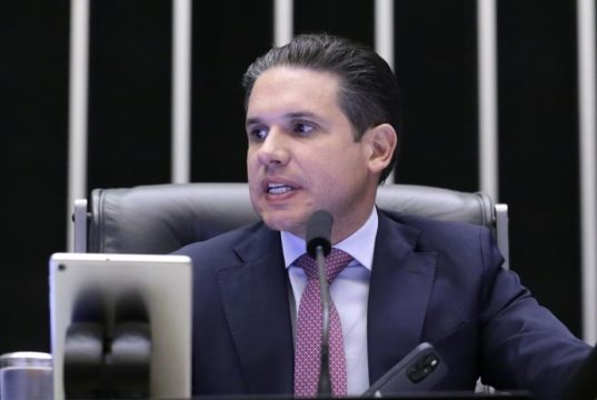 Hugo Motta olhando para o lado esquerdo e falando, sério, sentado, na mesa diretora da Câmara dos Deputados