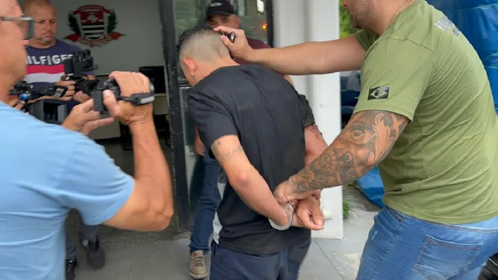 Douglas Alves da Silva chegando na delegacia, caminhando, de costas, de roupa preta, algemado com as mãos para trás