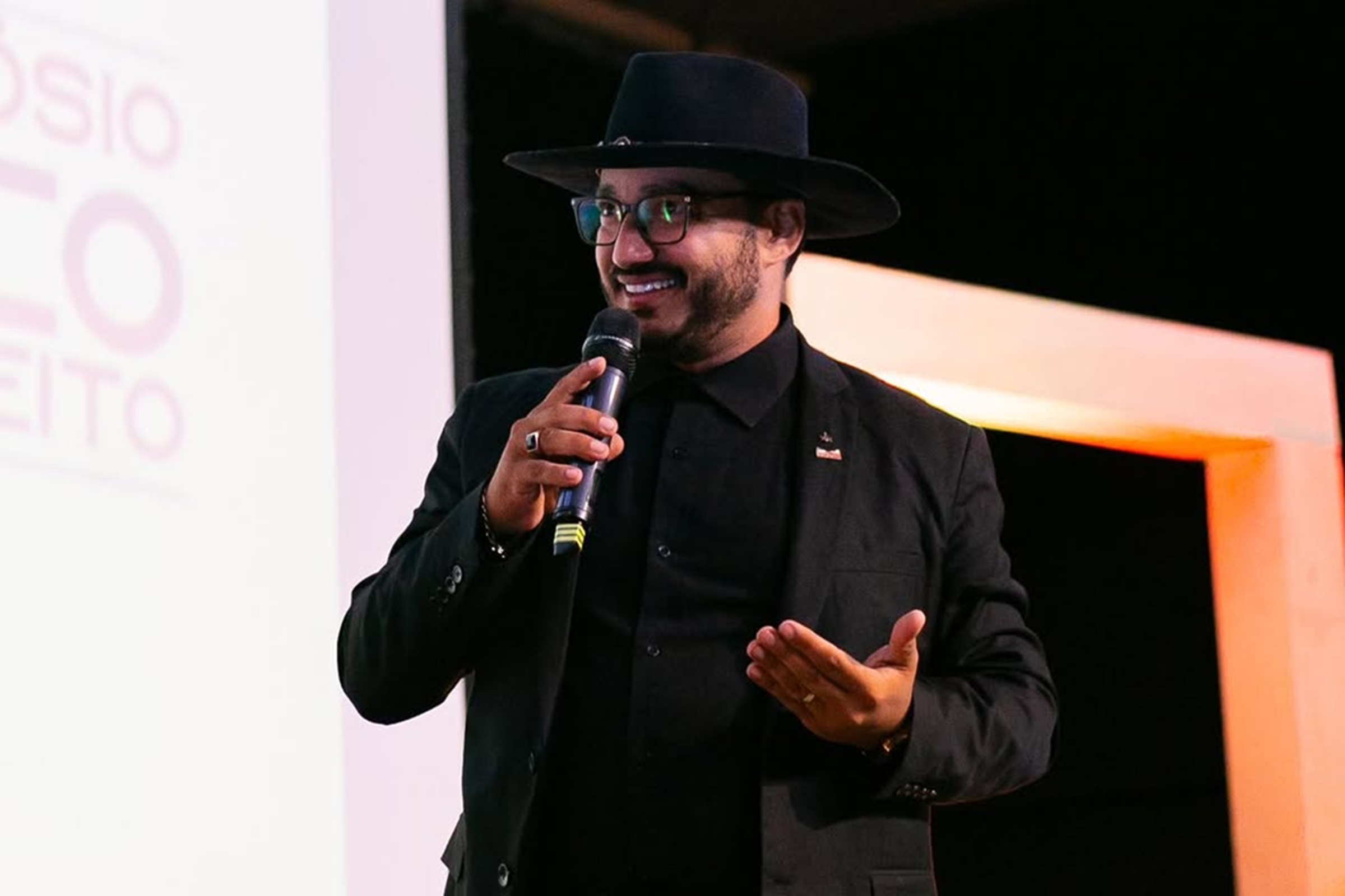 Dudu Morais, advogado, poeta e influenciador digital, falando e gesticulando em microfone, de roupa e chapéu pretos, olhando para o lado, sério