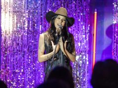 Ana Castela cantando em microfone, de roupa marrom e chapéu, de cabelo solto