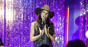 Ana Castela cantando em microfone, de roupa marrom e chapéu, de cabelo solto